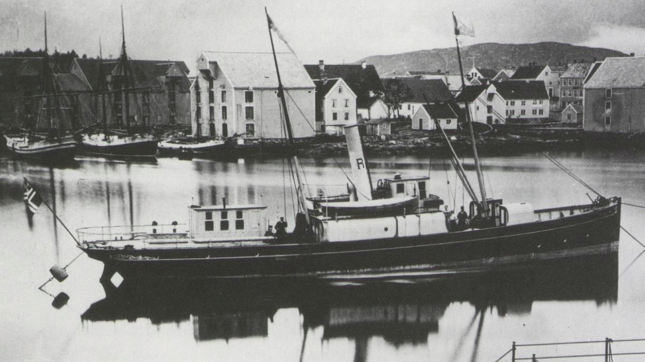 Historiskt svartvitt fotografi av ett ångfartyg förankrat i Stavangers hamn, med bebyggelse på andra sidan vattnet.